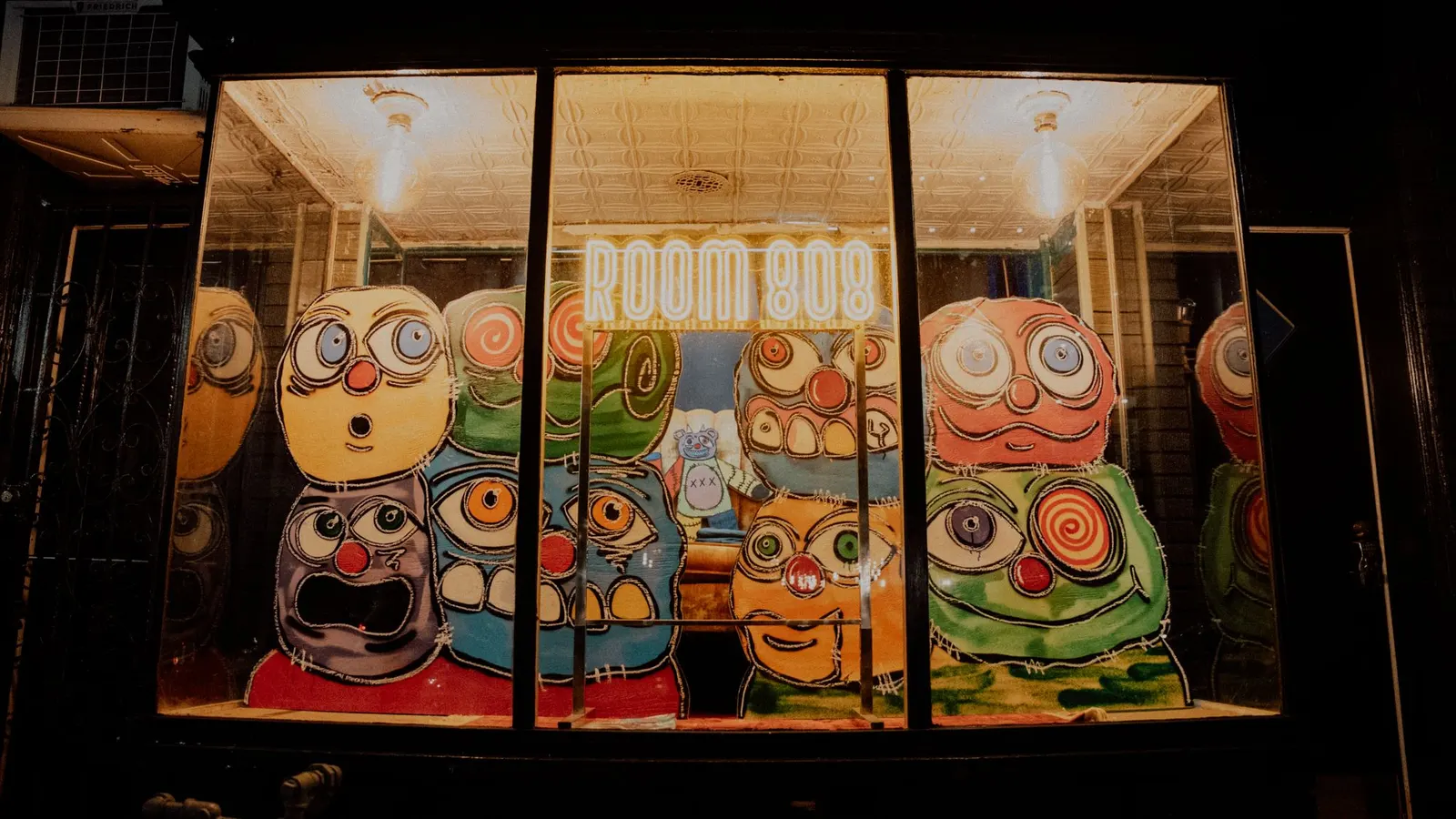 The Room 808 window at night: gold neon sign over painted faces